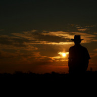 A farmer wearing a hat looks out over the Australian bush as the sunsets in the outback